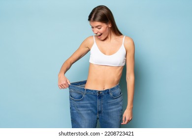 Woman Astonished With Results Of Her Slimming And Weight Loss Pulling Large Oversize Jeans To Check Her Success, Isolated On Blue Background With Wow Face Expression. Dieting, Keep Fit