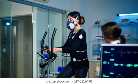 Woman athlete with mask running on cross trainer after medical researcher attached electrodes to her body. Physician using computer to controls EKG data showing on laboratory monitors, VO2 max test - Powered by Shutterstock