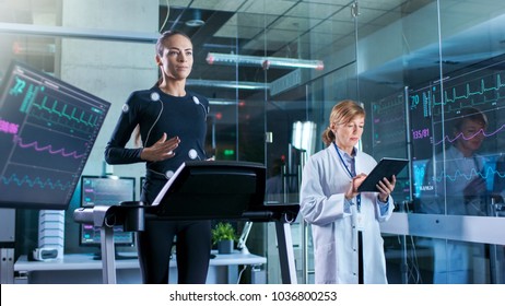 Woman Athlete Walks On A Treadmill  With Electrodes Attached To Her Body While Scientist Holding Tablet Computer Supervises Whole Process. In The Background Laboratory Monitors Showing EKG Readings.