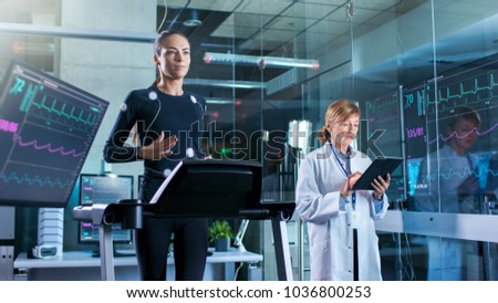 Woman Athlete Walks on a Treadmill  with Electrodes Attached to Her Body while Scientist Holding Tablet Computer Supervises whole Process. In the Background Laboratory Monitors Showing EKG Readings.