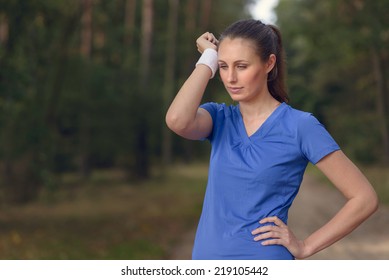 Woman Athlete Wiping Sweat From Her Forehead Onto Her Wristband As She Pauses During Her Training Exercises On A Forest Track