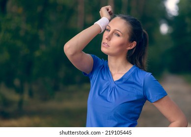 Woman Athlete Wiping Sweat From Her Forehead Onto Her Wristband As She Pauses During Her Training Exercises On A Forest Track