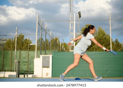 Woman in athletic attire playing badminton on an outdoor court. Green fencing and cloudy sky in the background. Active lifestyle and sport concept. - Powered by Shutterstock