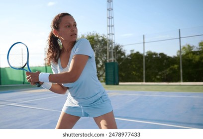 A woman in athletic attire playing tennis on an outdoor court. She looks focused and determined, holding a tennis racket in a ready position. - Powered by Shutterstock