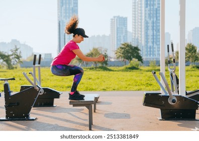 Woman in athletic wear performing a jump onto a platform at an outdoor gym, focused on her fitness routine, with a green park and buildings in the background - Powered by Shutterstock