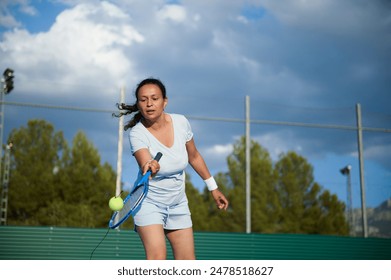 A woman in athletic wear playing tennis on an outdoor court beneath a cloudy sky. She is focused on hitting the tennis ball with her racket. - Powered by Shutterstock