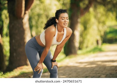 A woman in athletic wear is resting during her jog on a sunlit forest trail. She is leaning forward with hands on knees, wearing wireless earbuds, and looks relaxed in the peaceful surroundings. - Powered by Shutterstock