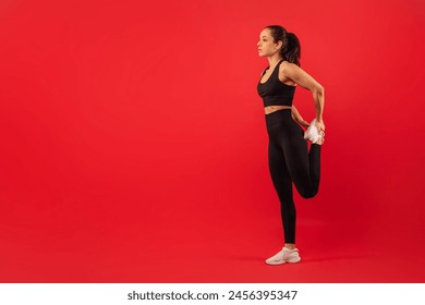 A woman in athletic wear is standing on one leg and holding her other foot behind her, stretching her quadriceps muscle. She maintains a focused expression, demonstrating proper form for a leg stretch - Powered by Shutterstock