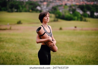 A woman in athletic wear stands in a grassy field, holding a rolled yoga mat over her shoulder, looking into the distance, ready for a yoga session in nature. - Powered by Shutterstock