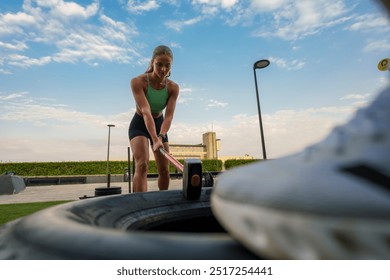 A woman in athletic wear is using a sledgehammer to hit a large tire outdoors. - Powered by Shutterstock