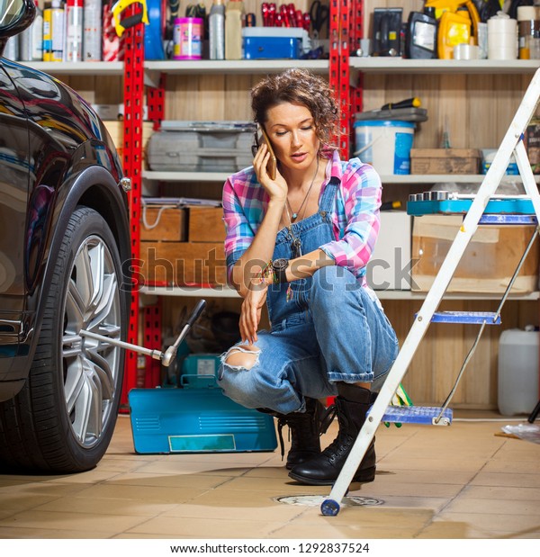 Woman auto\
mechanic in blue denim overals near the car in a car repair shop.\
Talking on the phone with the\
customer