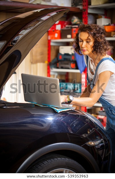 Woman auto mechanic uses a computer to diagnose\
car engine. Beautiful woman mechanic near a car with an open hood\
looking at a computer\
monitor