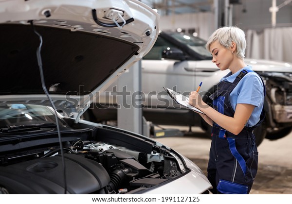 Woman auto\
Mechanic writing on clipboard at repair garage, wearing uniform\
overalls. Young mechanic engineer female taking notes on clipboard\
for examining a vehicle, side\
view