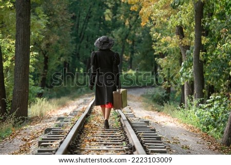 Woman in autumn fall forest, classic black coat and broad-brim hat walks along railway track with vintage suitcase. Back view.