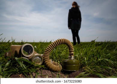 Woman Back To Camera Standing In Green Field On Blue Sky Background And Gas Mask Lying In Grass On Foreground. Enviromental Protection, Ecology, Earth Saving, Pollution Prevention And Hope Concept.