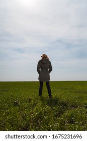 Woman Back To Camera Standing In Green Field On Clear Blue Sky Background And Gas Mask Lying In Grass On Foreground. Enviromental Protection, Ecology, Earth Saving, Pollution Prevention Concept.