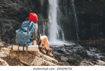 Woman with a backpack in a red hat dressed in active trekking clothes sitting near the mountain river waterfall and enjoying the splashing Nature power. Traveling, trekking, and a nature concept image - Shutterstock ID 2203290731