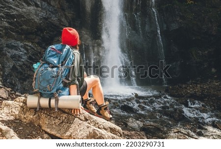 Woman with a backpack in a red hat dressed in active trekking clothes sitting near the mountain river waterfall and enjoying the splashing Nature power. Traveling, trekking, and a nature concept image