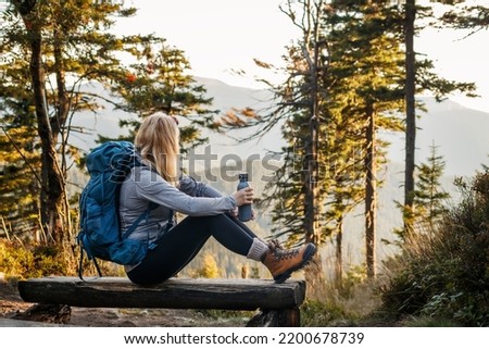 Woman with backpack and thermos resting on bench in forest. Relaxation during hiking in mountain