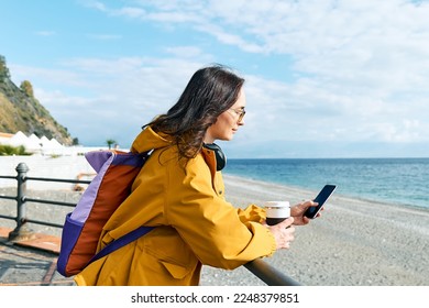 Woman with backpack using smartphone while leaning on handrail along seaside promenade. Relaxing outdoors. The concept of hiking and travel in the low season.