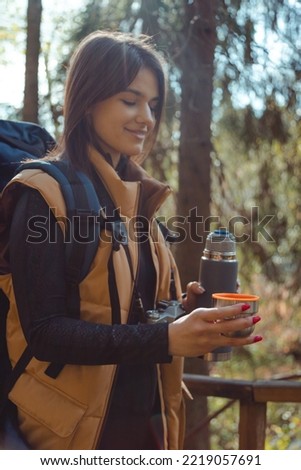 Woman backpacker holds thermos with hot beverage in hand at autumn forest, nature. Travel and camping concept. Warming drink