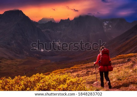 Woman Backpacking on Scenic Hiking Trail to Lake surrounded by Mountains during Fall in Canadian Nature. Colorful Dramatic Sunset Sky Artistic Render. Tombstone Territorial Park, Yukon, Canada.