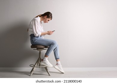Woman with bad posture using smartphone while sitting on stool near light grey wall indoors, space for text