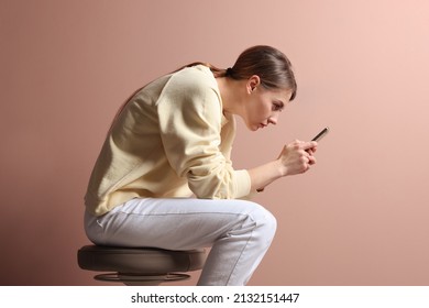 Woman with bad posture using smartphone while sitting on stool against pale pink background