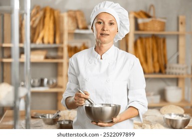 Woman baker cook stands against background of workspace with bowl and whisk in hands, posing, resting from work. Bakery worker picked up tool and got ready to bake buns - Powered by Shutterstock