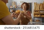 Woman baker handing over pastries to male customer in cozy bakery shop with shelves of breads in background