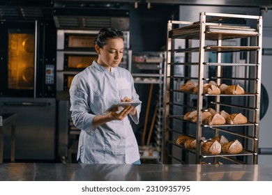 a woman baker in a uniform near the shelves with freshly baked bread holds tablet in her hands checks bread bakery - Powered by Shutterstock