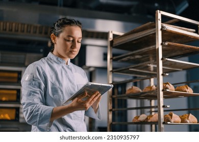 a woman baker in a uniform near the shelves with freshly baked bread holds tablet in her hands checks bread bakery - Powered by Shutterstock