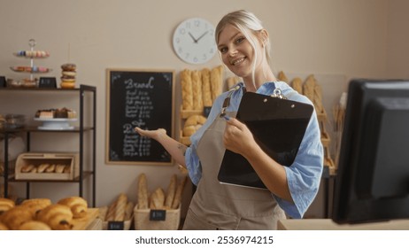 Woman in bakery holding clipboard smiling and pointing at chalkboard menu with fresh bread and pastries in background - Powered by Shutterstock