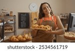 Woman in a bakery smiling while holding a tray of pastries, with breads in the background and a chalkboard menu on the wall