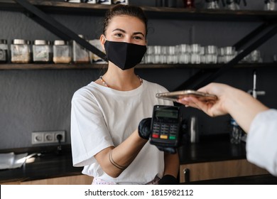 Woman Barista Wearing Medical Face Mask Holding Cashless Terminal To Pay For Order At The Counter, Customer Paying With Mobile Phone