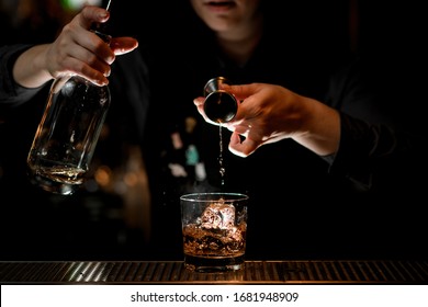 woman bartender elegantly pours alcoholic drink to glass with ice using beaker. Bottle with drink in her other hand. - Powered by Shutterstock