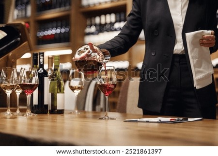 Woman bartender in strict classical clothes pouring red wine from decanter to the glass, close up image.