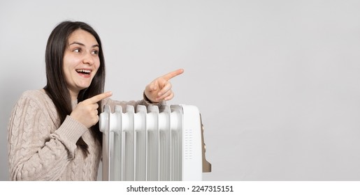A woman basks near an electric oil heater and points her fingers to the side in place for text. Methods of heating an apartment or house