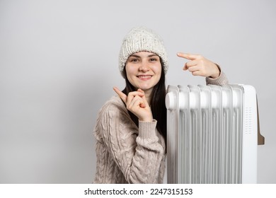 A woman basks near an electric oil heater and points her fingers to the side in place for text. Methods of heating an apartment or house