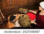 Woman in bath uniform doing whipped with broom steamed human male body in hot steam sauna, russian bath. Male lying relaxing on wood bench. Wellness, self-care, healthy concept. Copy ad text space