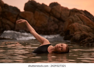 Woman bathes in sea at sunset, tranquil ocean swim, swimsuit-clad lady immerses for wellness, coastal rocks backdrop, peaceful water dip, eco-friendly leisure by seaside, health-centered retreat. - Powered by Shutterstock