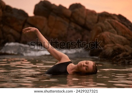 Woman bathes in sea at sunset, tranquil ocean swim, swimsuit-clad lady immerses for wellness, coastal rocks backdrop, peaceful water dip, eco-friendly leisure by seaside, health-centered retreat.