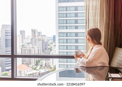 Woman in bathrobe with coffee cup sitting in modern hotel or apartment with city skyscraper view. Holidays in luxury hotel. Female person in hotel room on vacation