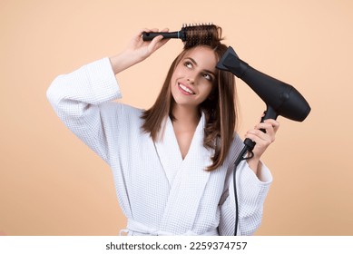 Woman in bathrobe combing hair, drying hair with hairdryer. Portrait of female model with a comb brushing hair. Girl with hairs brush and blow dryer. Hair care and beauty. Morning routine.