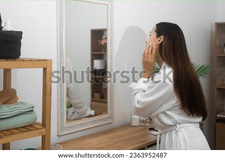 A woman in a bathrobe, at home, explores a beauty skincare concept, gazing into the bathroom mirror with care and intention. 