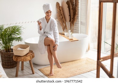 Woman In Bathrobe Moisturizing Smooth Legs With Moisturizer Cream Sitting On Bathtub In Modern Bathroom Indoors After Morning Shower. Beauty, Skincare And Pampering Concept