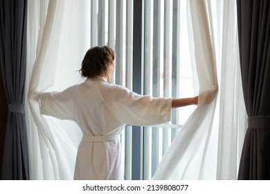 Woman In Bathrobe Opening Curtains And Looking Outside Through Window