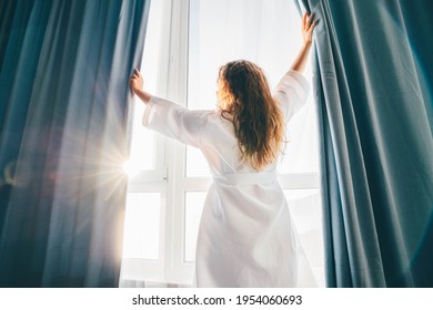 Woman in a bathrobe opening curtains in room at sunrise. 