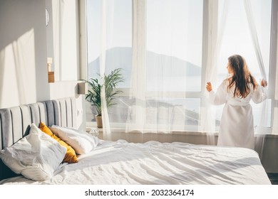 Woman in a bathrobe opening curtains in room at sunrise.  - Powered by Shutterstock