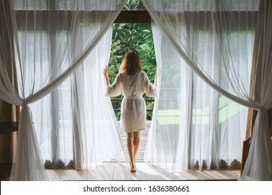 A woman in a bathrobe opens the curtains in deluxe Bali hotel room overlooking the terrace and tropical trees.Woman is awoke and standing before window. Girl is opening curtains and meeting sunrise - Powered by Shutterstock